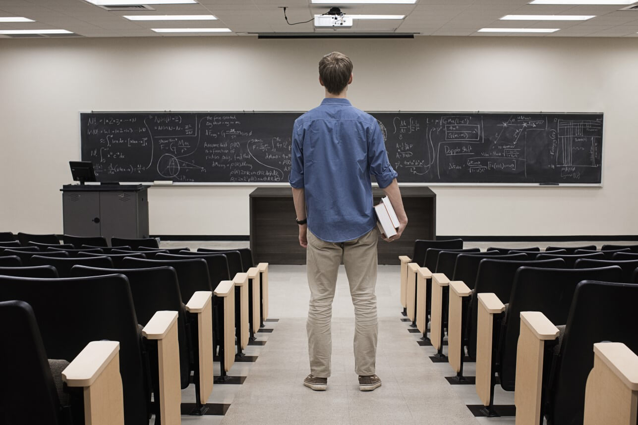 Student stands in the middle of an empty classroom, looking at a blackboard full of equations